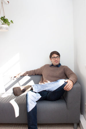 man on couch with crossed leg wearing blue jeans and handknit brown pullover