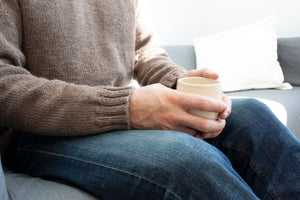close up of mans hands holding a white coffee cup, he is wearing blue jeans and handknit brown pullover
