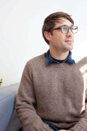 man sitting on grey couch wearing glasses and blue jeans modeling handknit raglan pullover